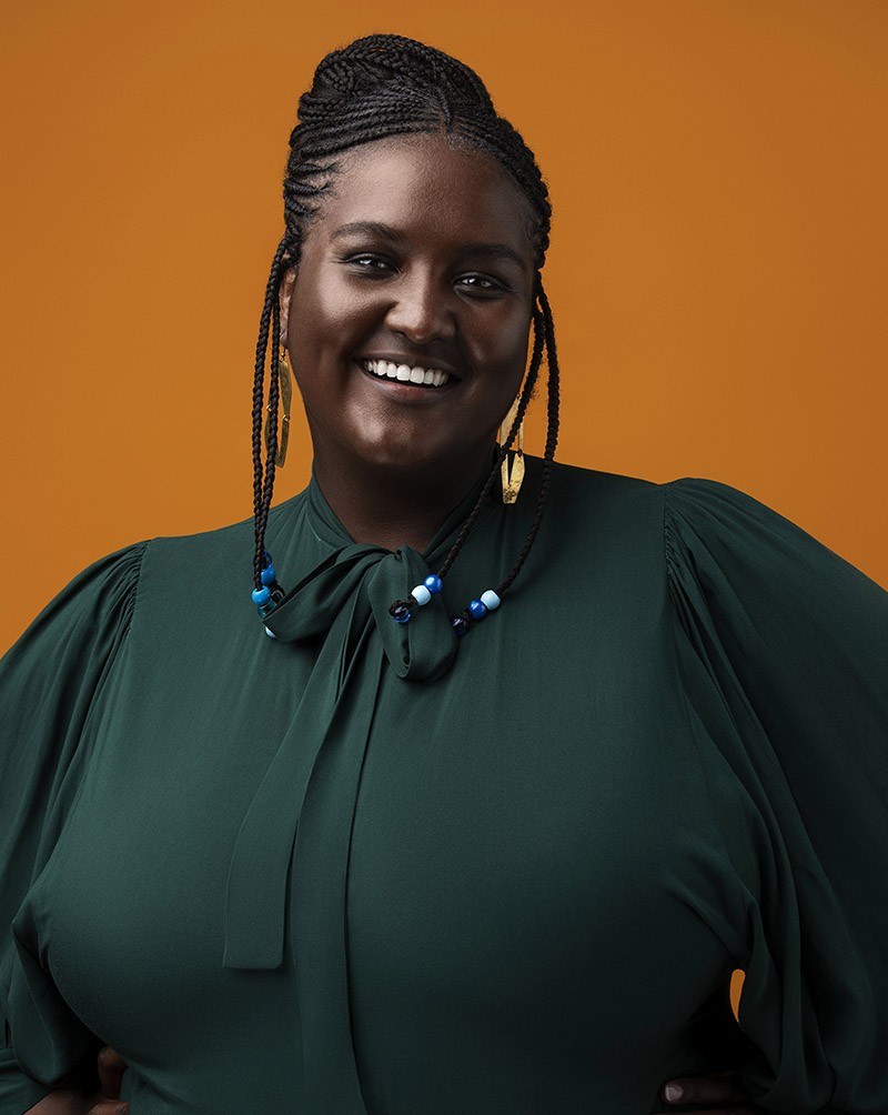 Sharrifa Ali in emeral green shirt with braided hair in front of an orange background.